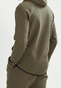 Olivgrön hoodie i mjukt tyg med hög krage, ribbade ärmslut och delad fåll bak. Har sidofickor och en strömlinjeformad design.