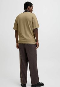 Khaki bomulds t-shirt med løs pasform, korte ærmer, båret med brune ternede bukser og sorte sko, set bagfra.