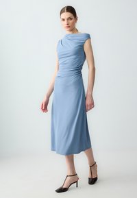 Abito azzurro chiaro realizzato in tessuto morbido, con scollatura drappeggiata, dettaglio arricciato sul lato e lunghezza midi, abbinato a scarpe col tacco nere.