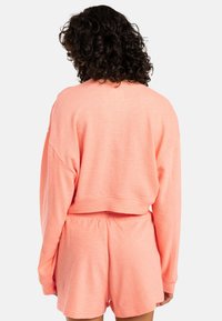 Perzikkleurig, kort sweatshirt met lange mouwen en een losse pasvorm, gecombineerd met bijpassende shorts met elastische taille. Zachte textuur, minimale stikdetails.