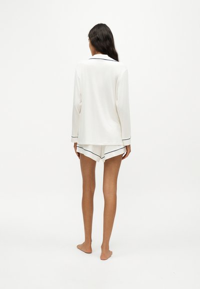 Femme debout pieds nus, de dos, portant un haut de pyjama blanc à manches longues et un short assorti avec une bordure sombre.