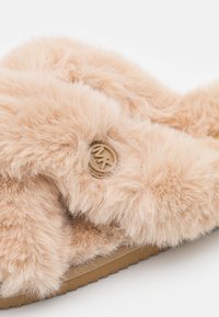 Gros plan d'une pantoufle beige et moelleuse avec des lanières croisées et un bouton rond avec logo sur le côté, dotée d'une semelle texturée.