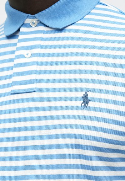 Κοντινό πλάνο ενός μπλουζάκι πόλο με ανοιχτό μπλε και λευκές ρίγες, δύο λευκά κουμπιά και ένα μικρό κεντημένο λογότυπο παίκτη πόλο.