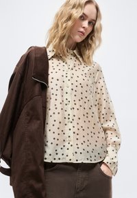 Młoda kobieta o falowanych blond włosach nosi beżową bluzkę w kropki i ciemnobrązową kurtkę zarzuconą na jedno ramię, z rękami w kieszeniach spodni.
