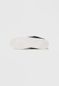 Černý teniska s horní částí z texturované látky a bílou gumovou podrážkou, s minimalistickým designem a vzorem podrážky ve tvaru rybí kosti.