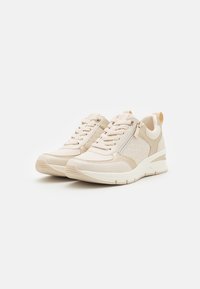 Beige sneakers med en teksturert side, hvit såle, snøring foran og en glidelåsdetalj på siden. Glatt materiale og rund tådesign.
