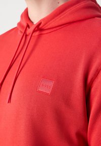 Gros plan d'une personne portant un sweat à capuche rouge avec des cordons et un petit logo carré embossé indiquant "BOSS" sur la poitrine.