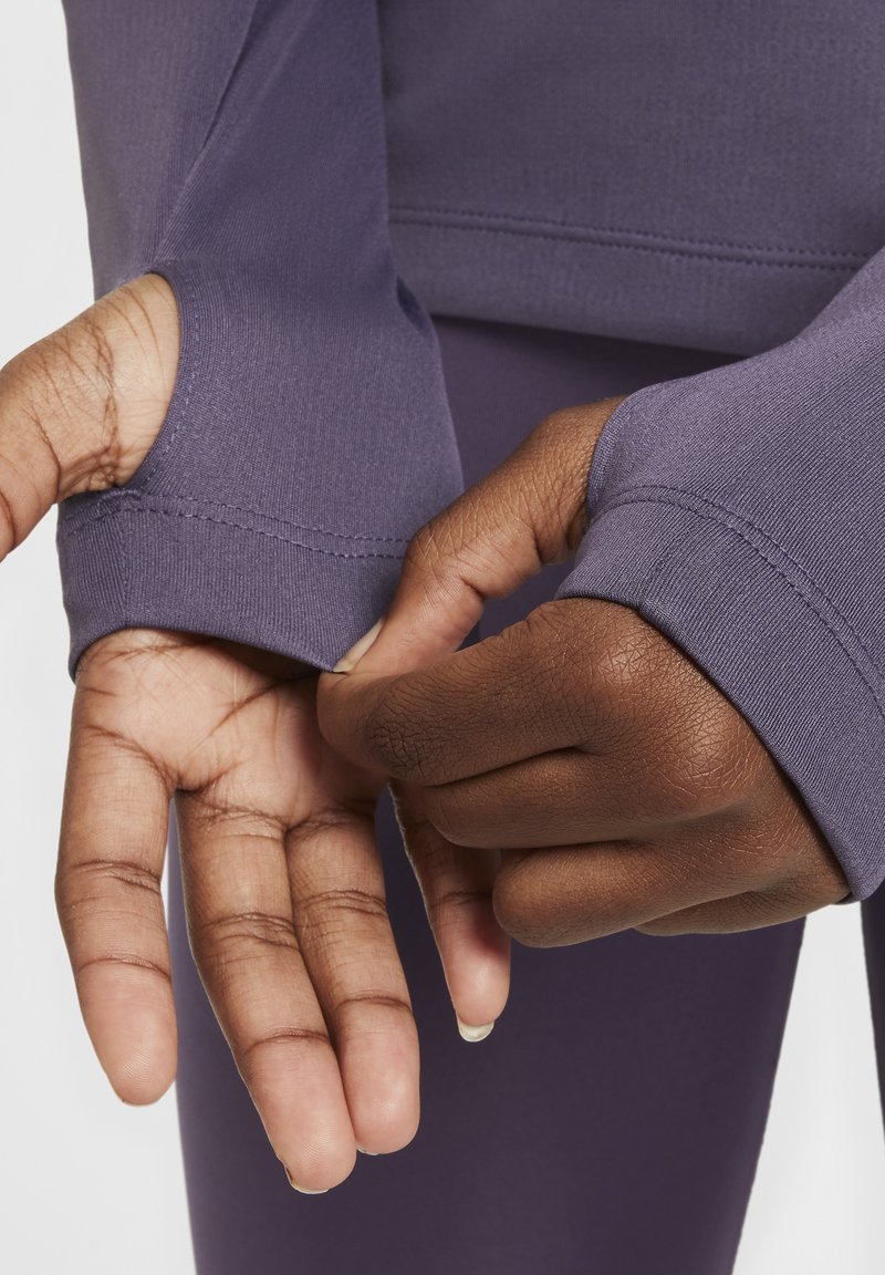 Top lungo viola con maniche, dettaglio del foro per il pollice, mostrato mentre le mani regolano il polsino, enfatizzando il tessuto morbido e il design aderente.