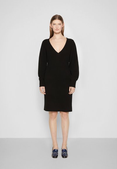 Vestido preto de malha com decote em V, mangas longas e silhueta ajustada. Combinado com sapatos de bloco azul-marinho com detalhes metálicos.