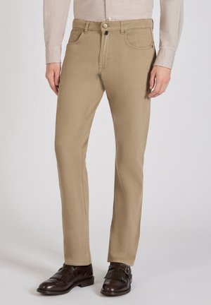 Chino kaki avec un design à jambe droite, en tissu coton et avec des coutures visibles. Associé à des chaussures de ville marron foncé.