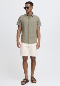 Camisa de lino verde oliva de manga corta, pantalones cortos crema con vuelta, gafas de sol oscuras y sandalias marrones sobre un fondo claro.