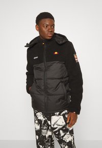 Schwarze isolierte Jacke mit einem Fleece-Oberteil, Reißverschluss und farbenfrohen Logo-Details auf der Brust. Auffälliger, gepolsterter unterer Teil und Kapuze.