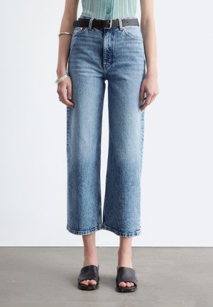 Person iført lyseblå ribbet ærmeløs top, højtsiddende brede cropped blå jeans, sort bælte, sorte åbne tæer sandaler og sølv armbånd.