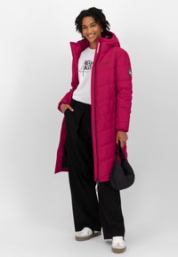 Langer magentafarbener Puffer-Mantel mit Kapuze, Frontreißverschluss und seitlichen Taschen, getragen über einem weißen Shirt und schwarzen weiten Hosen. Schwarze Handtasche inklusive.