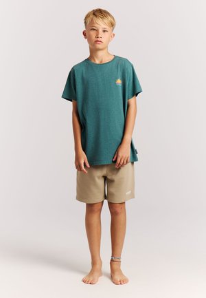 Blond jongetje staat op blote voeten, draagt een teal T-shirt met zonafbeelding en beige korte broek tegen een effen lichte achtergrond.
