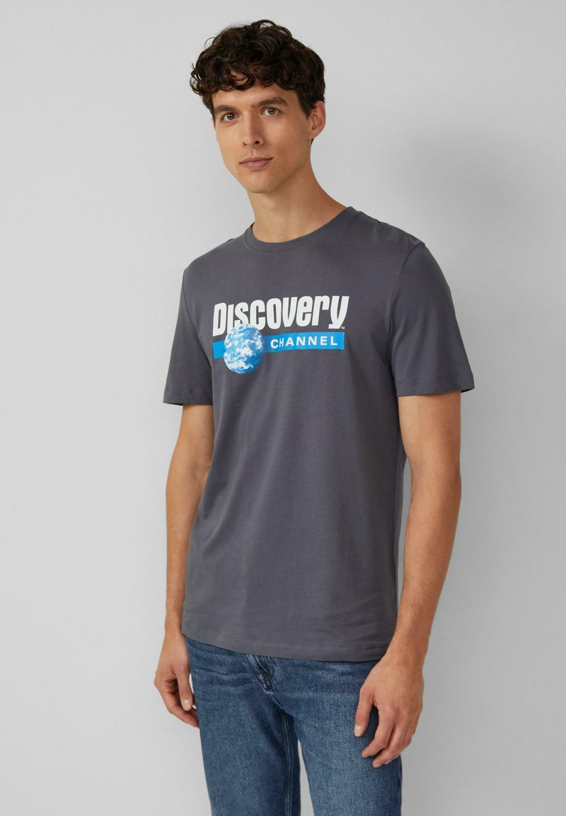 Grijze katoenen T-shirt met het "Discovery Channel" logo in wit en blauw, voorzien van een wereldbolafbeelding aan de linkerkant en korte mouwen.