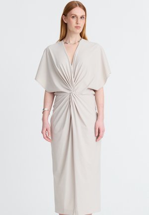 Frau trägt ein hellbeiges Kleid mit weiten Ärmeln und einem verdrehten Knotendetail an der Taille und steht vor einem einfachen weißen Hintergrund.
