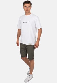 Witte katoenen t-shirt met zwart scriptlogo, gecombineerd met olijfgroene shorts. Sneakers zijn wit met rode accenten. Casual outfit weergegeven.