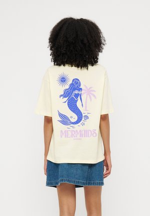 Femme aux cheveux bouclés portant une chemise crème montrant une sirène bleue, le soleil, un palmier, des coquillages et le texte "MERMAIDS" dans le dos.