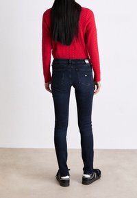 Jeans ajustados de mezclilla oscura, con una cintura baja, dos bolsillos traseros y una cinturilla con marca. Combinados con un suéter de punto rojo y zapatos negros.