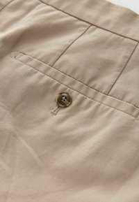 Tela de algodón beige con un pequeño bolsillo trasero y un botón redondo de color marrón oscuro. Superficie texturizada con sutiles líneas diagonales.
