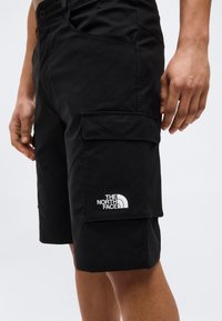 Μαύρα cargo σορτς με πλαϊνή θήκη και το λογότυπο The North Face που φοριούνται από ένα άτομο που στέκεται με τα χέρια χαλαρά.