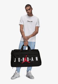 Sac de sport noir avec le grand texte blanc "JORDAN" et le logo Jumpman rouge. Dispose de deux poignées et d'un dessus zippé, fabriqué à partir de matériaux durables.