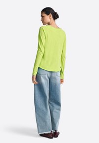 Dlhý rukáv, svetlo limetkovo zelený top z mäkkej tkaniny, kombinovaný so svetlomodrými vysoko pásovými nohavicami s širokými nohavicami a tmavými, textúrovanými topánkami.