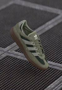 Olíva zöld sneaker nubuk és hálós anyagból, három fehér csíkkal, fűzős elülső résszel és texturált barna gumitalppal.