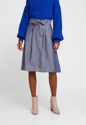 Kvinde iført en blå ribstrikket sweater, knælængde grå nederdel med bundet bælte og beige ankelstøvler, stående mod en hvid baggrund.