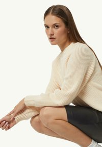 Cremefarbener gerippter Pullover mit hohem Kragen und langen Ärmeln, kombiniert mit einem schwarzen Lederrock, der die strukturierte Stoffqualität und die lässige Passform zeigt.
