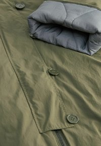 Veste vert olive avec fermeture par boutons et fermeture éclair, avec une doublure intérieure matelassée grise pliée au niveau du poignet.