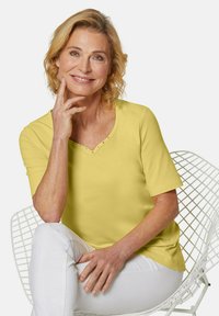 Gelbes kurzärmliges Oberteil mit V-Ausschnitt und dekorativen Nieten am Ausschnitt, kombiniert mit weißen Hosen. Glatte Stoffstruktur.