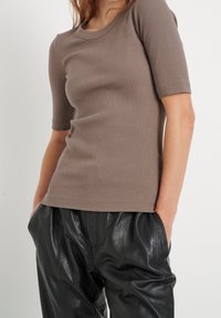 Brązowa, prążkowana bluzka z krótkim rękawem i okrągłym dekoltem, połączona z czarnymi skórzanymi spodniami. Posiada małe logo na dolnym brzegu.