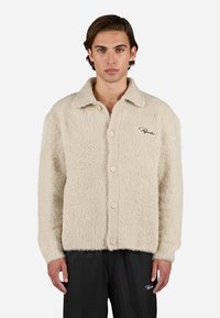 Beige, fluffy knap-shirt med spidse kraver og lange ærmer. Har fire knapper og et diskret broderet logo på brystet.