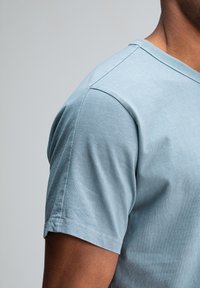 Close-up van een persoon die een lichtblauwe katoenen t-shirt met korte mouwen draagt, waarbij de schouder en bovenarm zichtbaar zijn tegen een effen achtergrond.