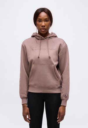 Kvinde står vendt fremad iført en malva Champion hoodie med frontlomme og sorte bukser mod en ensfarvet grå baggrund.