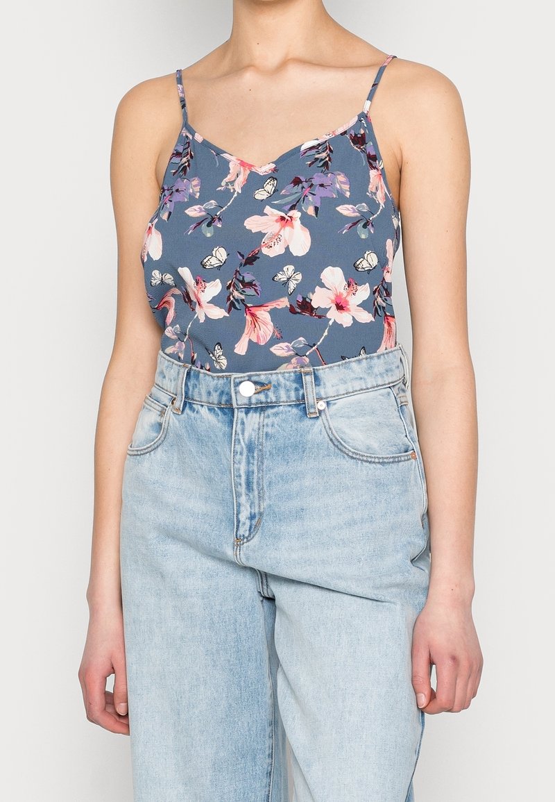 Femme portant un débardeur bleu à bretelles fines à motifs floraux, rentré dans un jean clair taille haute, debout devant un fond uni.