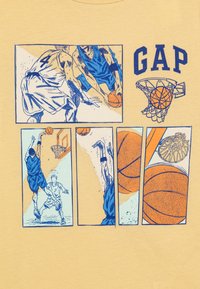 T-shirt jaune clair avec des illustrations de baskets bleues et oranges, un logo "GAP" et des panneaux de style bande dessinée représentant des joueurs et des ballons de basket.
