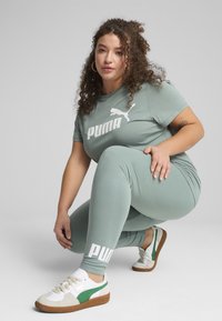 Světle zelené vypasované tričko a legíny s bílým logem "PUMA". Model má na sobě bílé tenisky se zelenými detaily a hnědými podrážkami.