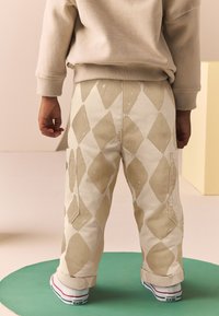 Beiget collegehousut, joissa on suuri timantinmuotoinen kuvio ja sivutaskut, yhdistettyinä valkoisiin lenkkareihin, joissa on punaisia ja sinisiä yksityiskohtia.