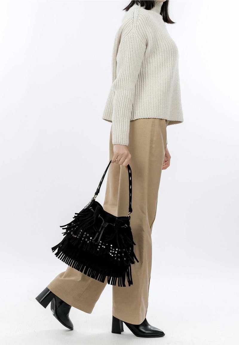 Femme portant un pantalon beige à jambes larges, un pull en maille crème, des bottes à talons noires, tenant un sac à main noir à franges avec des clous dans la main droite.