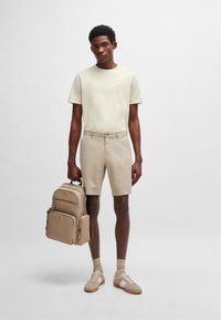 Camiseta de algodón beige, pantalones cortos beige claros y mochila beige con múltiples compartimentos. Zapatos color crema con detalles en blanco, de tela texturizada.