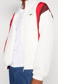 Biała męska kurtka wiatrówka z czerwonymi i brązowymi panelami, zamkiem błyskawicznym, elastycznymi mankietami oraz małym zielonym logo na piersi.