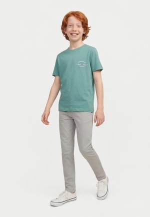 Vörös hajú fiú mosolyog, türkiz színű mintás pólót, világosszürke nadrágot és fehér tornacipőt visel, fehér háttér előtt sétál.