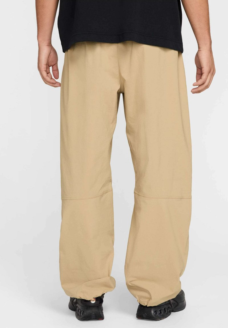 Khakifarbene weit geschnittene Hose mit lockerer Passform, ausgestattet mit einer glatten Textur und gesteppten Akzenten, getragen mit schwarzem Schuhwerk.