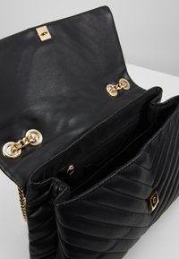 Černá prošívaná kožená kabelka se zlatým řetízkovým popruhem, otevřeným klopovým uzávěrem, vnitřní zipovou kapsou a zlatým zapínáním na bílé ploše.