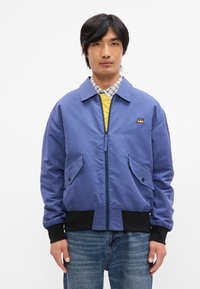 Μπλε ελαφρύ μπουφάν τύπου bomber με μπροστινό φερμουάρ, ελαφρώς ελαστικές μανσέτες και δύο μπροστινές τσέπες. Διαθέτει μια μικρή κεντημένη λεπτομέρεια σε σχήμα κορώνας.