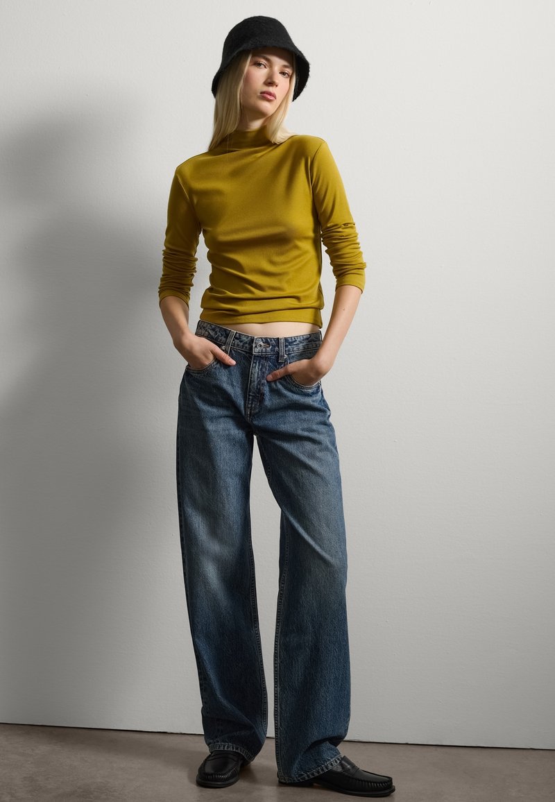 Top jaune moutarde à manches longues avec taille cintrée, associé à un jean en denim bleu ample et un chapeau seau noir. Le mannequin se tient avec les mains dans les poches.