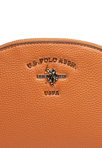 Oranžová texturovaná kožená taška s logem U.S. Polo Assn. ve zlaté barvě a zipovým detailem na vrchu.